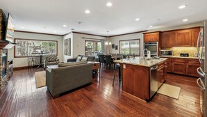 Een ruime woonkamer met een open haard, een bank, een eettafel en een keuken met houten kasten en roestvrijstalen apparatuur.