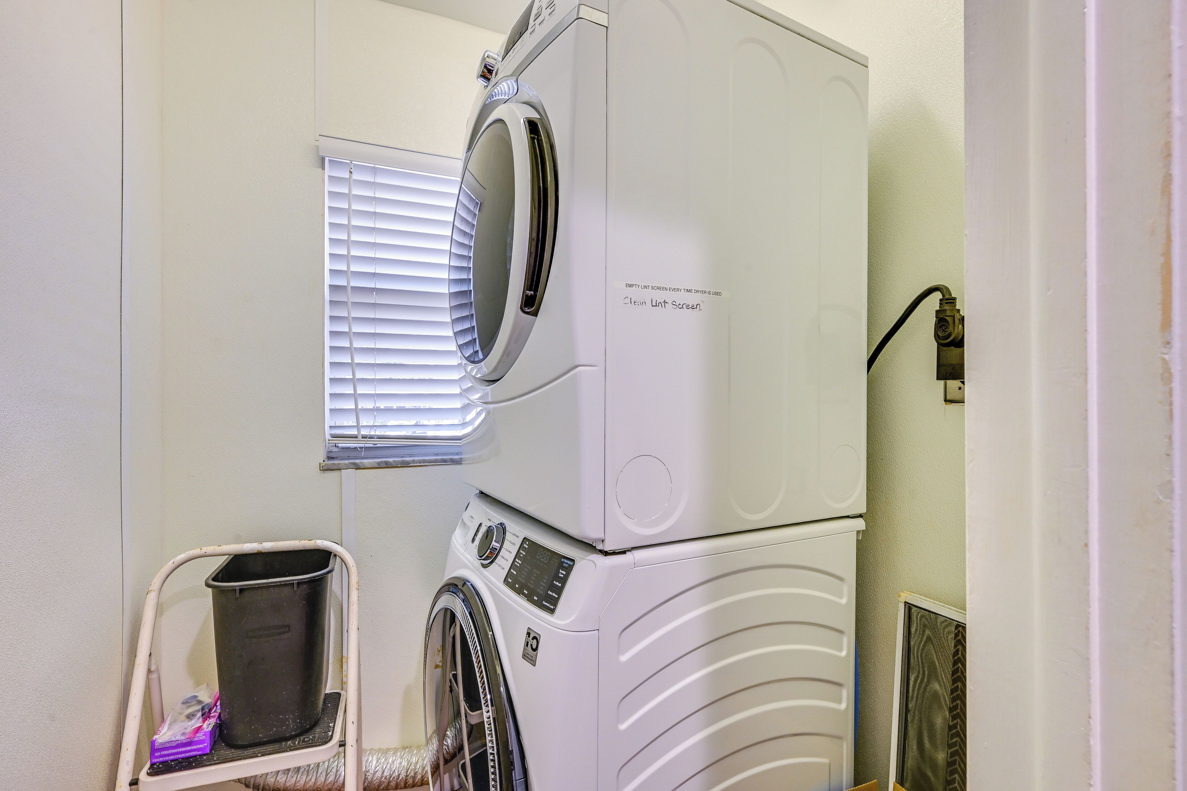 A stacked washer and dryer unit with a control panel, a window with blinds, and a metal laundry basket.