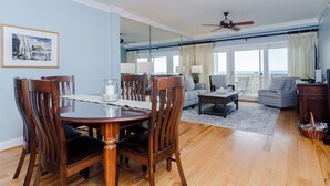 A living room with a dining area, a ceiling fan, a large window with curtains, and a view of the outdoors. - Upgraded Oceanfront 3BR Condo in WrightsvilleDunes (Wrightsville Beach)