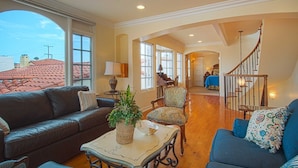 A spacious living room with a leather sofa, a patterned armchair, and a coffee table with a plant. A staircase leads to a bedroom with a blue bedspread. - Beautiful, bright and charming 3-story townhouse on an amazing street in Newport! (Newport Beach)