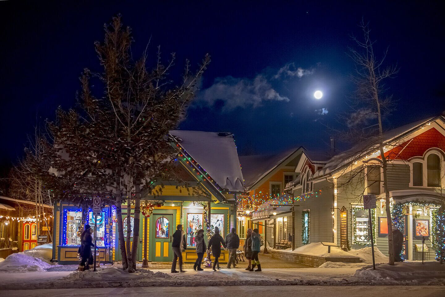 A snow-covered street at night with illuminated buildings and a visible moon.