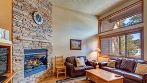 Una sala de estar con chimenea de piedra, sofá de cuero, mesa de centro de madera y ventanas amplias.