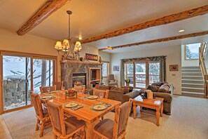 A spacious living area with a fireplace, wooden beams, large windows, and a staircase leading to a balcony. - Rustic Home on Whitefish Mountain, Near Ski Run! (Whitefish)