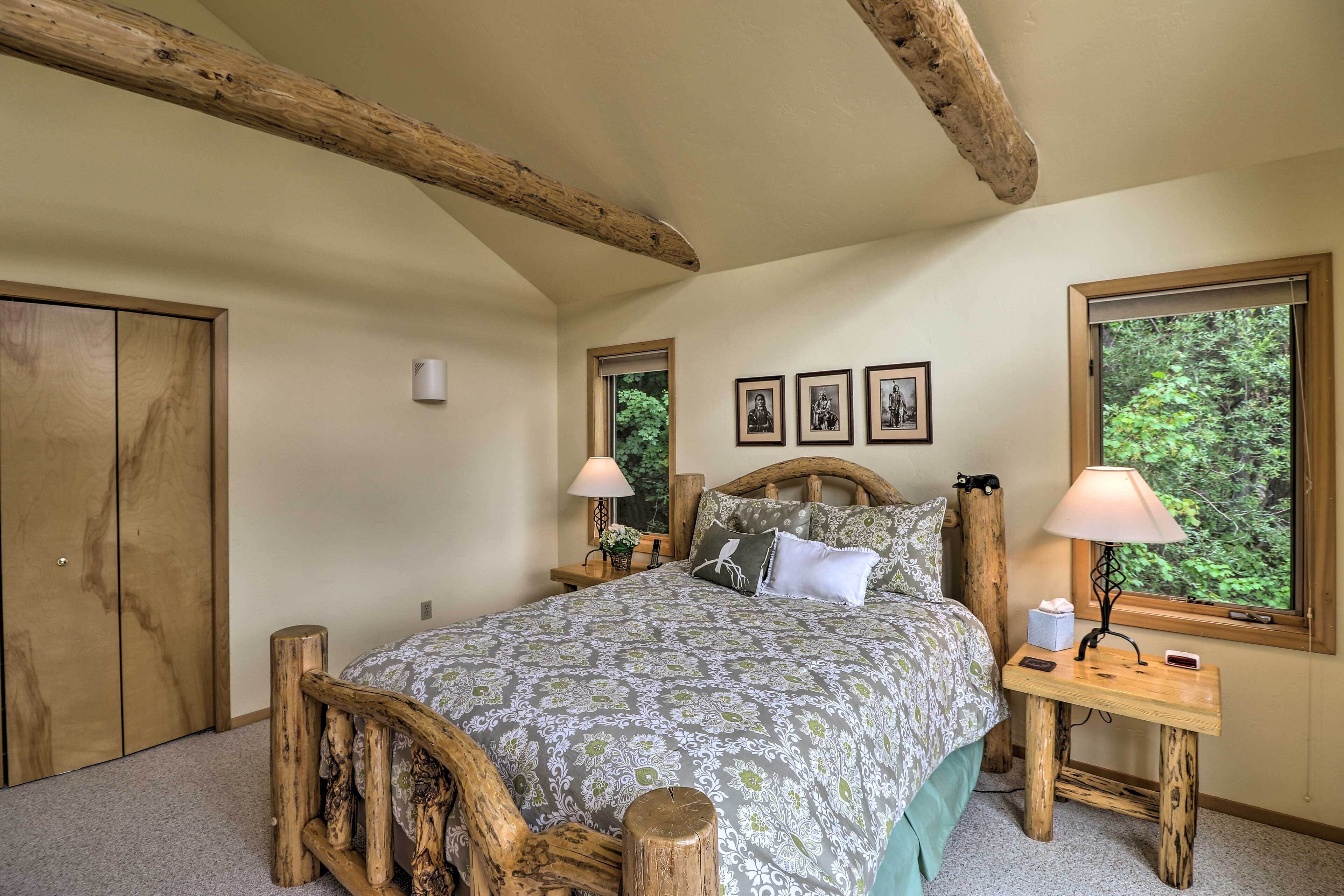 Een slaapkamer met een houten bedframe, een nachtkastje met een lamp en een raam met uitzicht op bomen.