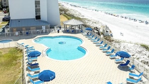 A pool area with lounge chairs and umbrellas, a beach in the background, and a tall building nearby. - Beach House Condominium 202B by Newman-Dailey (Miramar Beach)