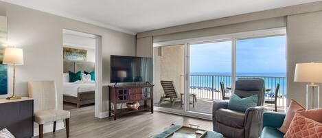 A spacious living room with a large window overlooking the beach, a comfortable sofa, a television, and a dining area.