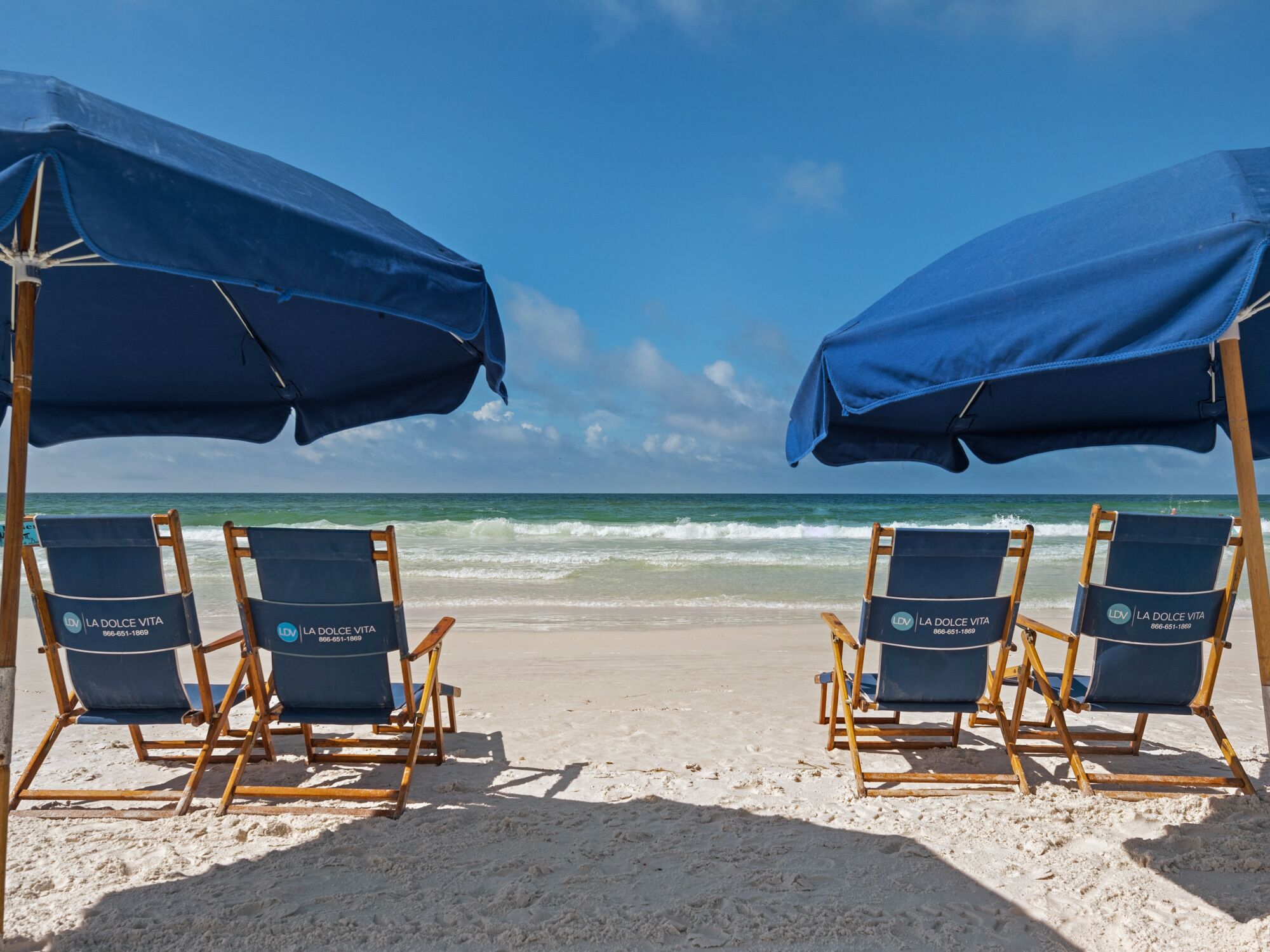 Two rows of beach chairs under blue umbrellas on a sandy beach with the ocean in the background.