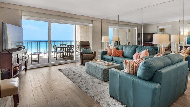 A modern living room with a large sofa, a television, and a balcony with a view of the ocean. - Beach House Condominium 202B by Newman-Dailey (Miramar Beach)