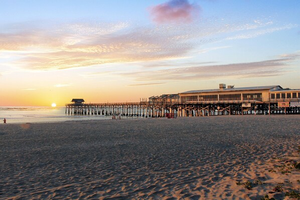 A beach with a pier extending into the water, a building with a flat roof, and the sun setting in the background.