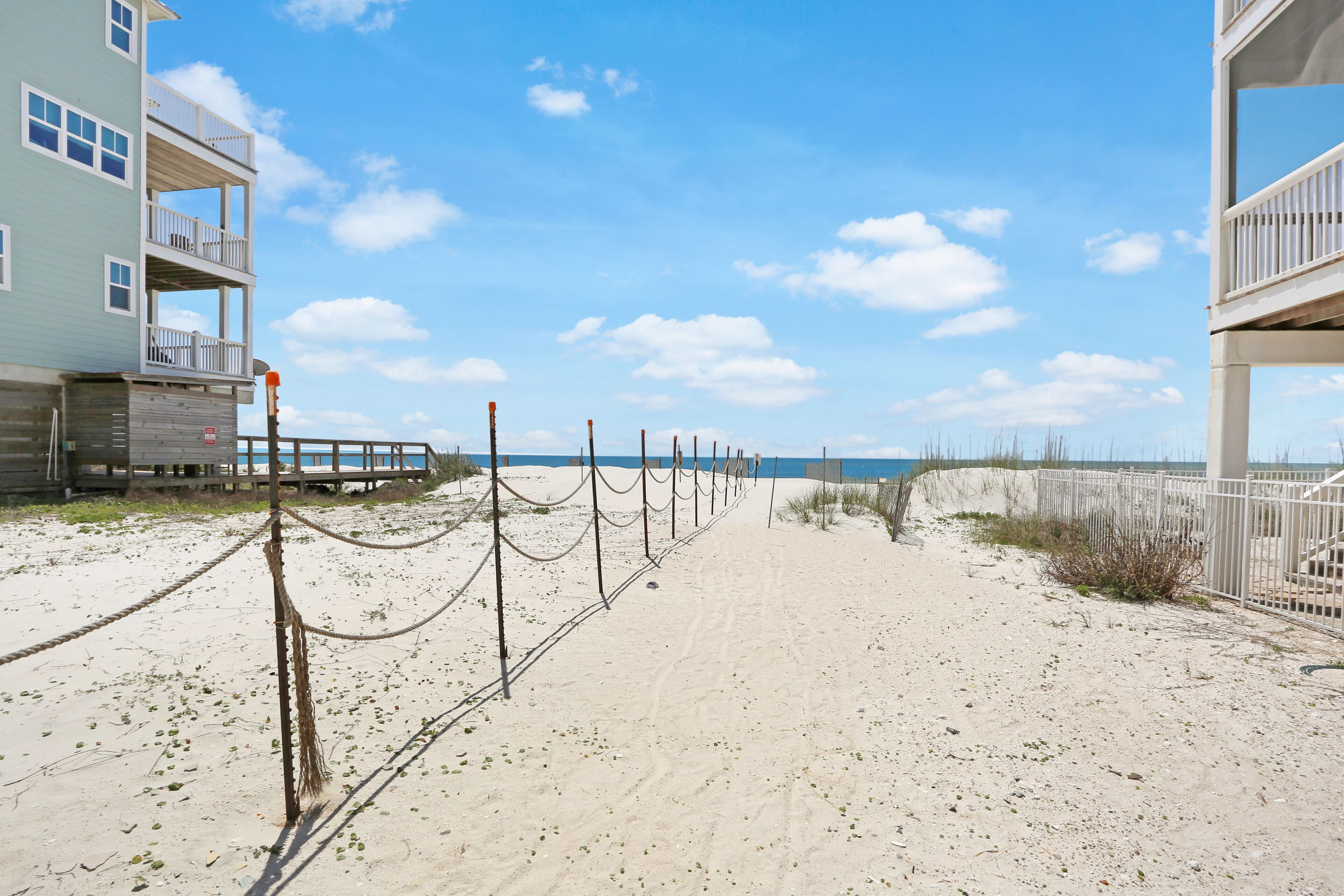 A sandy beach path with a wooden fence and a building with balconies.