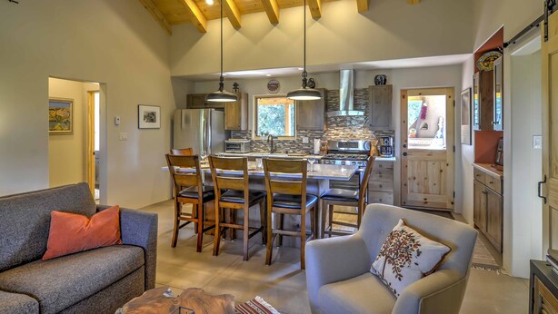 A spacious living area with a kitchen in the background, wooden ceiling beams, and a dining area with chairs and a table. - Hidden Santa Fe Gem - Nestled in Tesuque Hills! (Santa Fe)