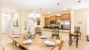 A dining area with a wooden table set for four, surrounded by chairs, a chandelier, and a kitchen in the background. - Storey Lake Resort home with Mini-Golf & Lazy River | Private Pool | Sleeps 10 (Kissimmee)