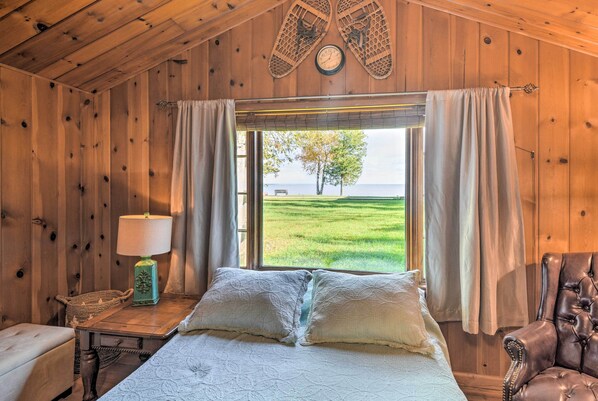 A wooden cabin bedroom with a bed, a nightstand, a lamp, and a view of a grassy field.