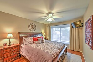 A bedroom with a bed, nightstands, a ceiling fan, a TV, a window with curtains, and a decorative wall piece. - Hot Tub & Pool: Stunning Goodyear Home Near Lakes (Goodyear)
