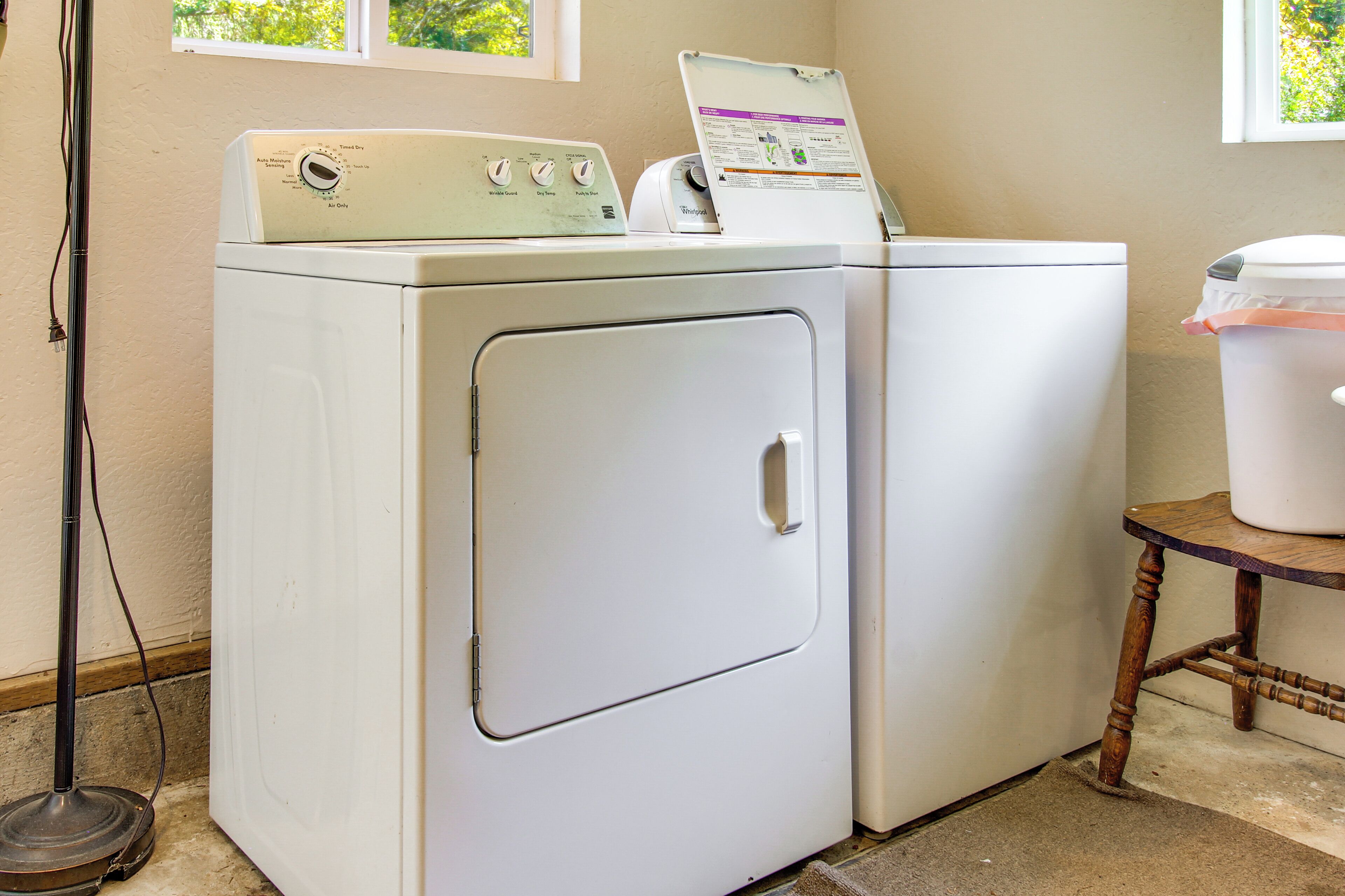 A laundry area with two white washing machines, a wooden table with a container, and a window with a view of greenery.