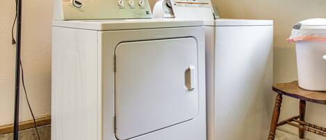 A laundry area with two white washing machines, a wooden table with a container, and a window with a view of greenery.