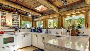 A kitchen with wooden beams, white cabinets, a large window, and a countertop with various appliances. - Hike to the Coast! Log Cabin on 5 Acres in Bandon (Bandon)
