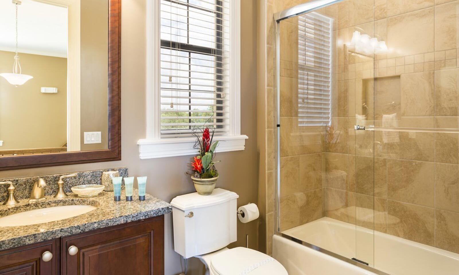 A bathroom with a granite countertop, a toilet, a shower with glass doors, and a window with blinds.