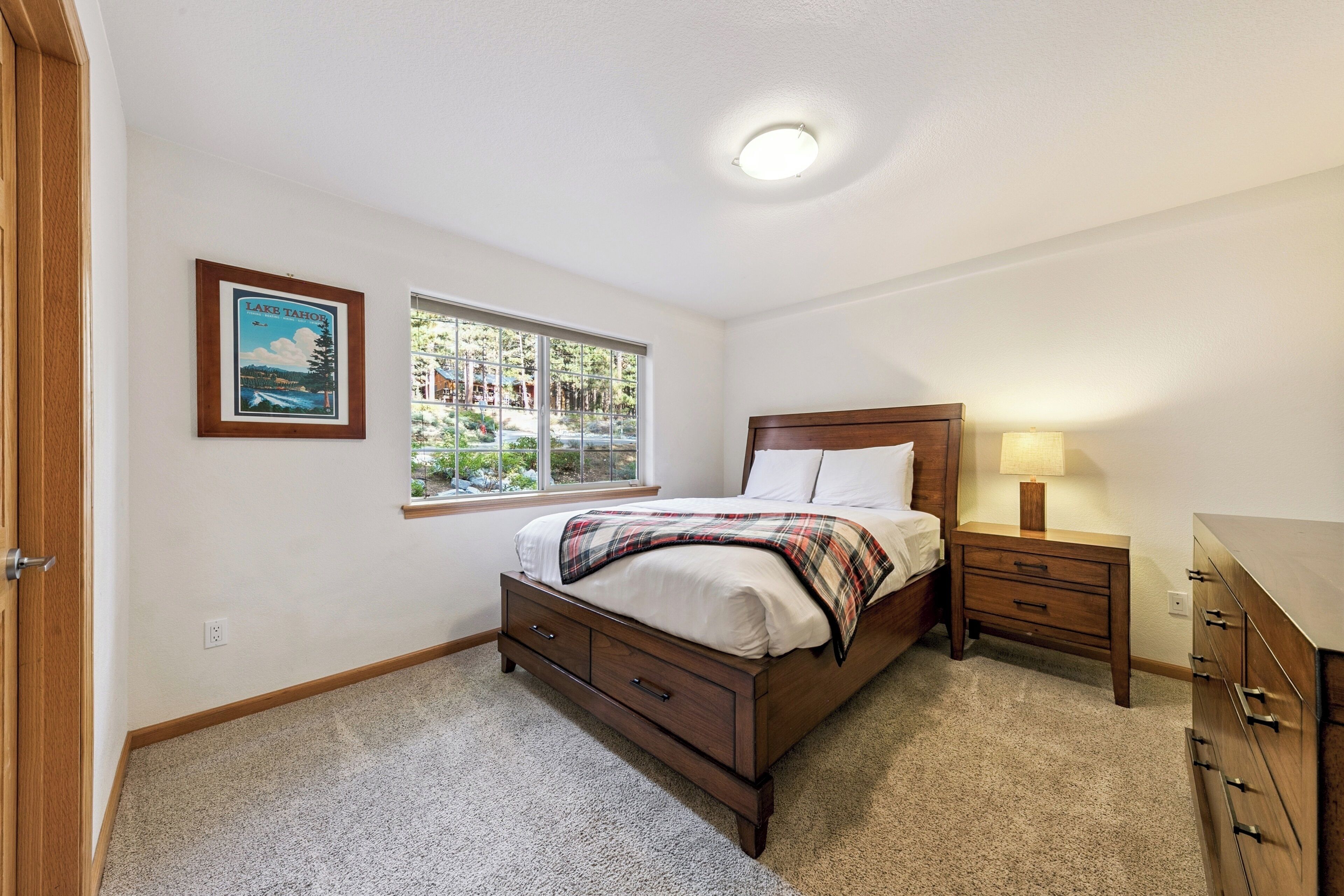 A bedroom with a bed, nightstand, dresser, and a window with a view of trees.