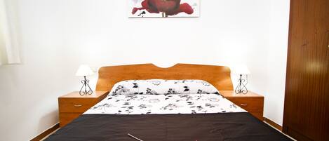 Una cama bien hecha con una colcha negra, dos lámparas de noche, una mesita de noche de madera y un cuadro de una flor roja colgado en la pared.