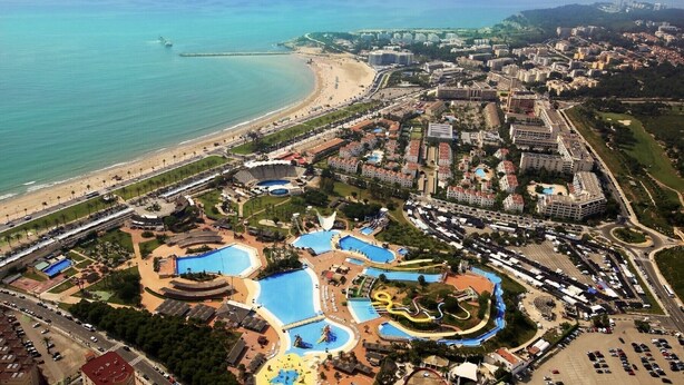 Vista aérea de un complejo turístico costero con una gran piscina, playa y un desarrollo urbano denso al fondo.