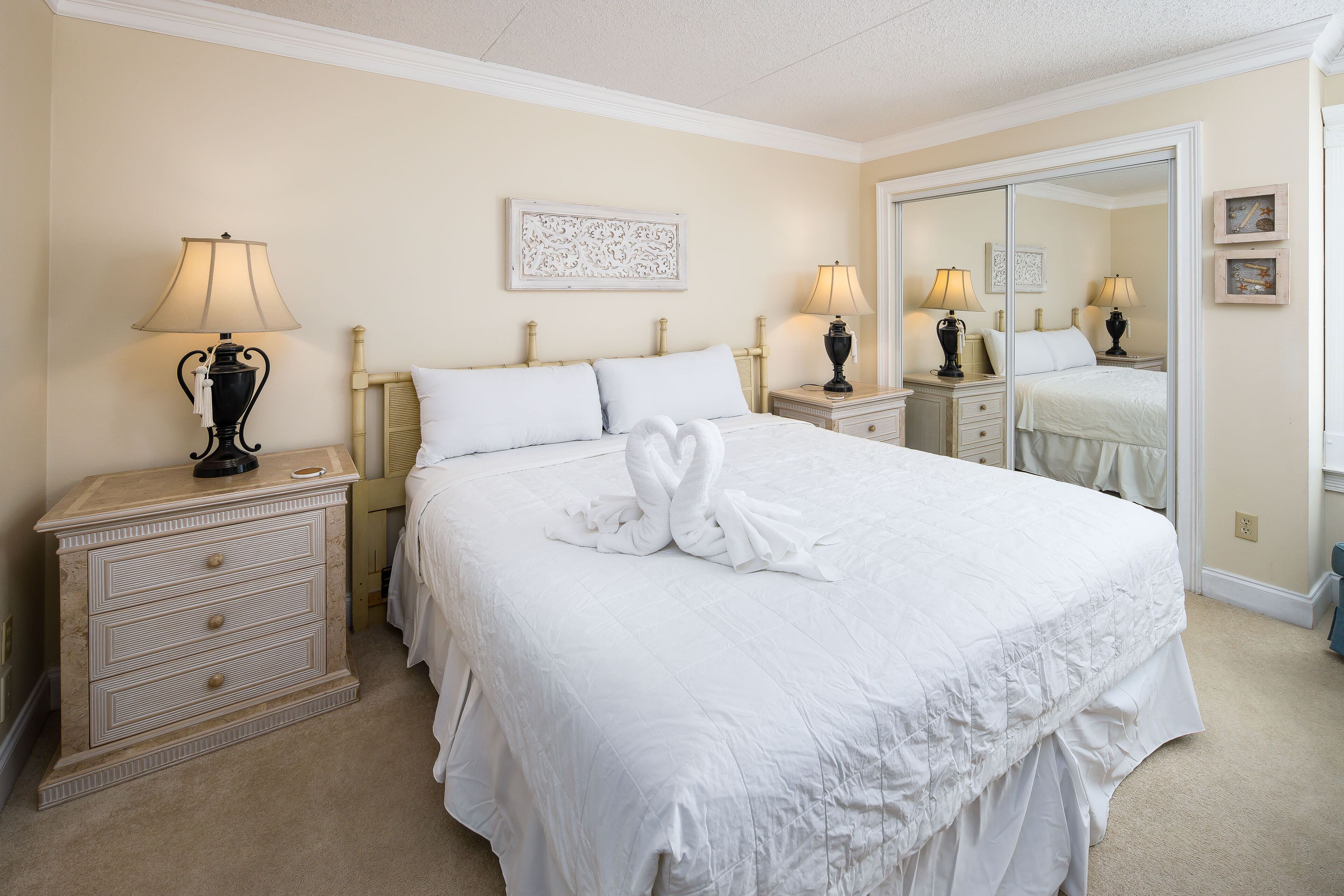 A neatly made bed with a towel swan, a bedside lamp, a nightstand, a dresser, and a mirror.