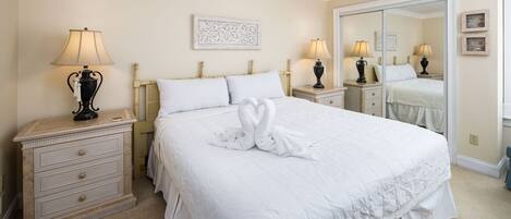 A neatly made bed with a towel swan, a bedside lamp, a nightstand, a dresser, and a mirror.