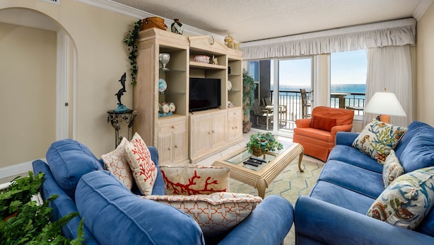 A living room with blue sofas, a wooden entertainment unit, and a view of the sea through large windows.