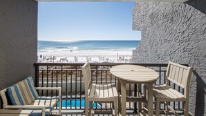 A balcony with a view of the beach, a pool, and wooden furniture.