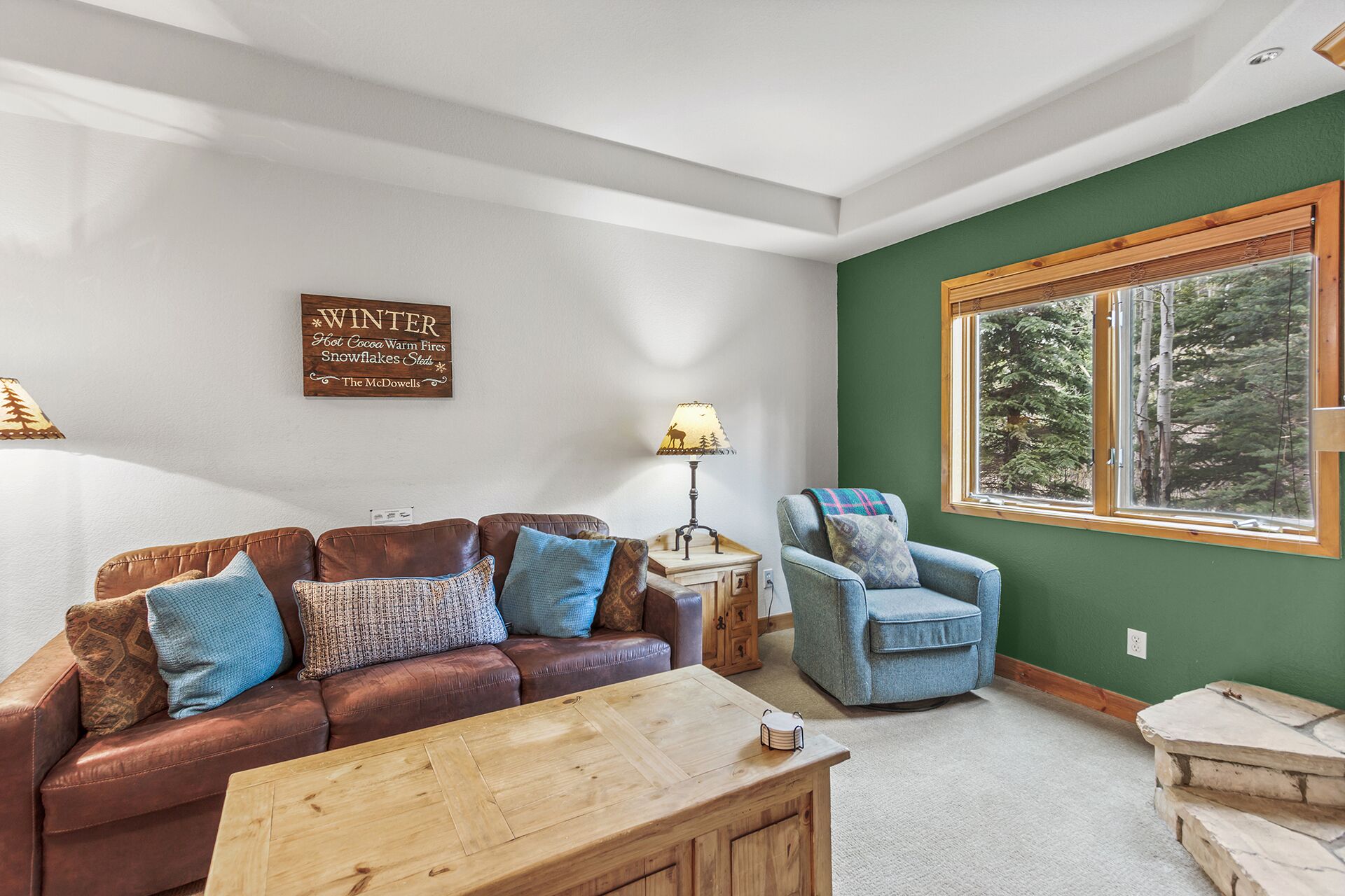 A cozy living room with a brown leather sofa, a blue armchair, and a wooden coffee table. A sign on the wall reads 'WINTER Hot Cocoa Warm Fires Snowflakes Skids.'