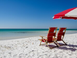 Eine Strandansicht mit einem roten und weißen Strandkorb und zwei hölzernen Liegestühlen im Sand.