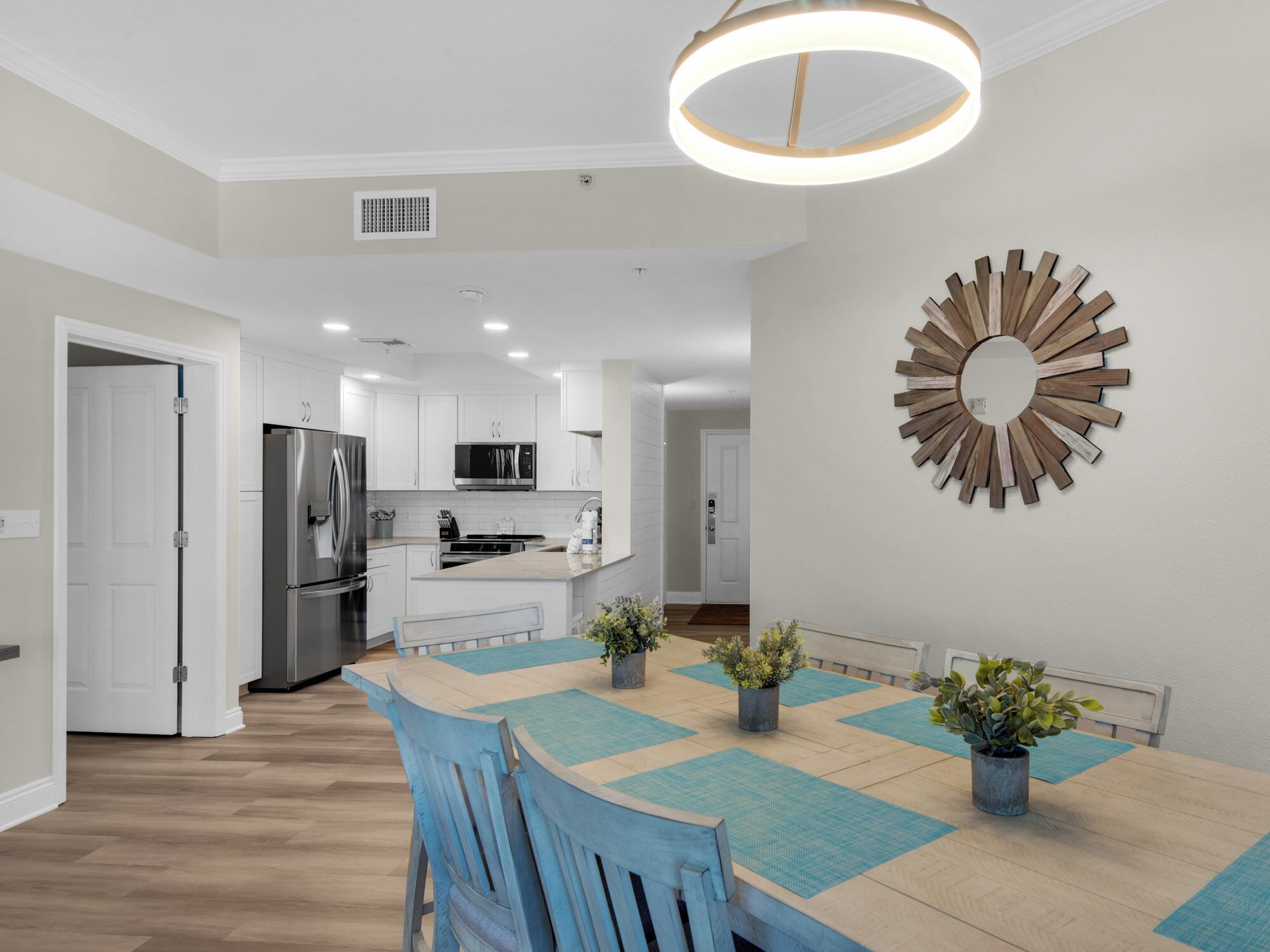Une cuisine moderne dotée d’appareils électroménagers en acier inoxydable, un coin repas avec une table en bois et des sets de table bleus, ainsi qu’un élément décoratif en bois fixé au mur.