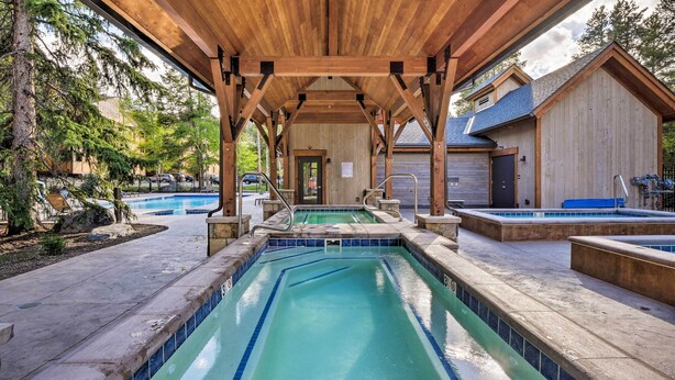 Zona de piscina techada con vigas de madera, un jacuzzi independiente y vista a un edificio al fondo.