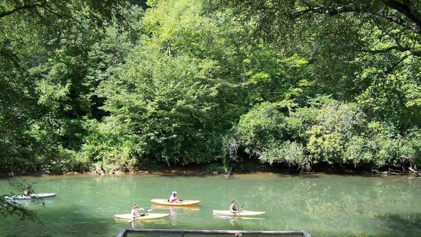 Terraza de madera con asientos exteriores y vista a un río con kayaks.