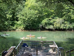 A wooden deck with outdoor seating and a view of a river with kayaks.