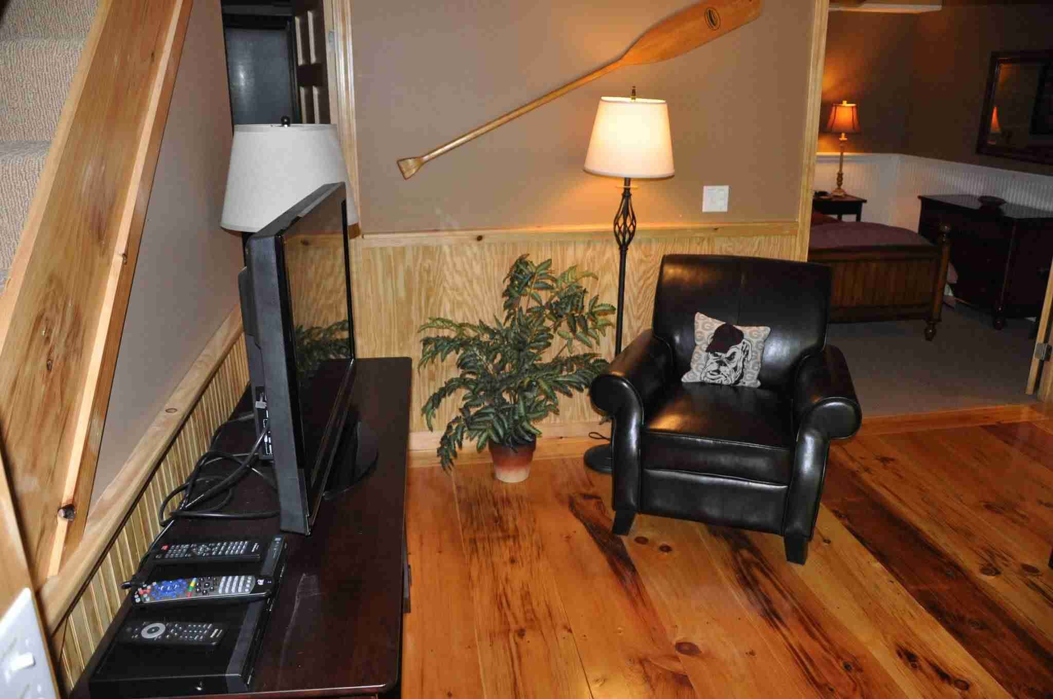 Een kamer met een houten vloer, een zwarte leren fauteuil, een lamp, een plant en een houten wand met latten.