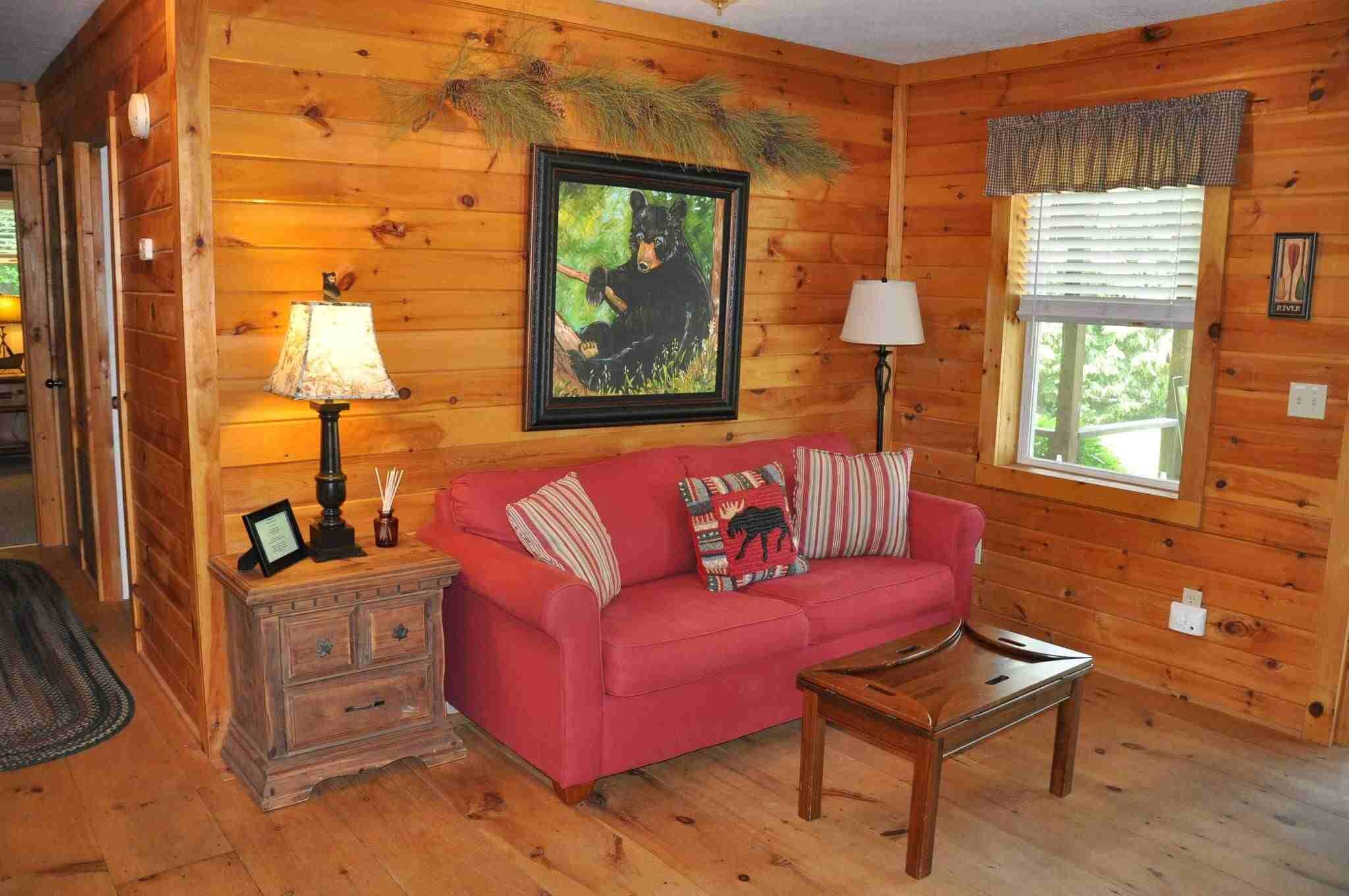 L’intérieur chaleureux d’un chalet avec un mur en bois, un tableau représentant un ours encadré, un canapé rouge orné de coussins à motifs d’élans, une table d’appoint en bois avec une lampe, et une fenêtre avec des stores.