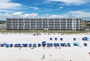 A multi-story hotel with a flat roof, numerous balconies, and a beach area with umbrellas and lounge chairs.