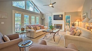 A spacious living room with a high ceiling, large windows, a fireplace, and a television. - Charming Hilton Head Home on Quiet Street (Hilton Head Island)