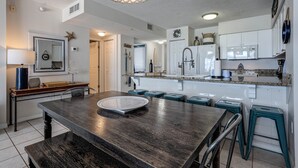A modern kitchen with white cabinets, a granite countertop, and a dining area with a dark wooden table and teal bar stools. - Island Princess 609 * Gulf front~Beach Chair Set Included * Salt Water Vacations (Fort Walton Beach)