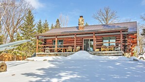 A log cabin with a stone chimney, wooden porch, and snow-covered ground. - Wonderful Home on Sand Lake w/ Expansive Porch! (Spring Lake)