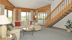 A spacious living room with a high ceiling, a wooden staircase, and large windows. - WH974- Huge Private Pool, only 500 Feet To Beach, Basketball, & Dog Friendly! (Corolla)