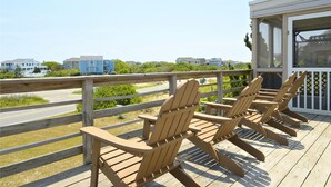 A wooden deck with Adirondack chairs and a covered seating area overlooking a road and residential buildings. - WH974- Huge Private Pool, only 500 Feet To Beach, Basketball, & Dog Friendly! (Corolla)