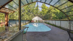 Piscina exterior techada con techo curvo, rodeada de árboles y un patio con sillas y una mesa.