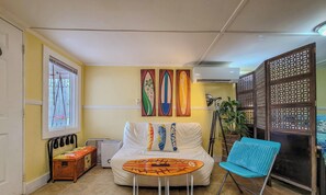 A room with a bed, a chair, a small table, a window, and a wall with surfboard art.