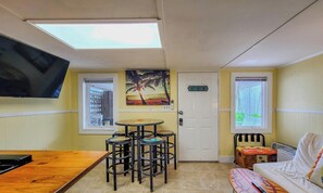 A compact room with a wooden table, three stools, a TV mounted on the wall, a door, and two windows.