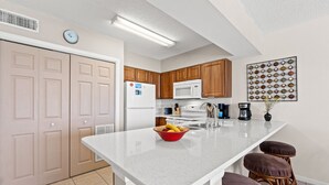 A kitchen with white cabinets, a white refrigerator, a microwave, a sink, and a countertop with a bowl of fruit. There are wooden stools and a clock on the wall.