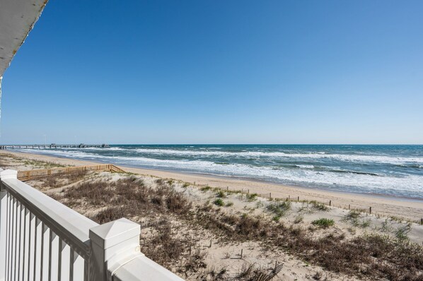 Vista de una playa con un paseo de madera que conduce al agua, dunas de arena y un cielo azul despejado.