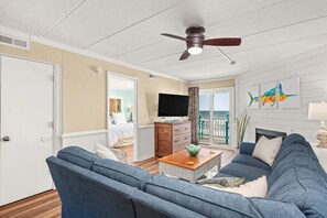 A living room with a blue sofa, a wooden coffee table, a ceiling fan, and a TV. - The Paisley Marlin: PierView Oceanfront Condo, Cozy Amenities and Incredible Vie (Nags Head)