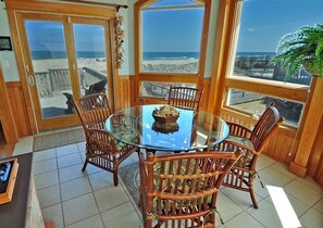 Relax in this serene sunroom with a glass table and wicker chairs, surrounded by expansive windows overlooking the beach.